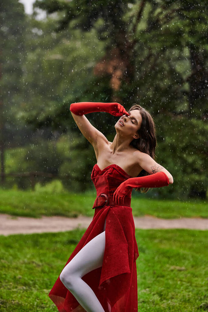 Μια νεαρή γυναίκα με ένα κόκκινο φόρεμα και μακριά γάντια στέκεται με χάρη στη βροχή, αγκαλιάζοντας το καλοκαιρινό αεράκι στη φύση. - Φωτογραφία, εικόνα