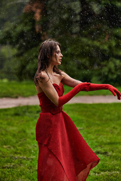 Dalgalı kırmızı elbiseli ve uzun eldivenli genç bir kadın yaz yağmurunda zarif bir şekilde dans ediyor. Duruş ve zarafeti simgeliyor.. - Fotoğraf, Görsel