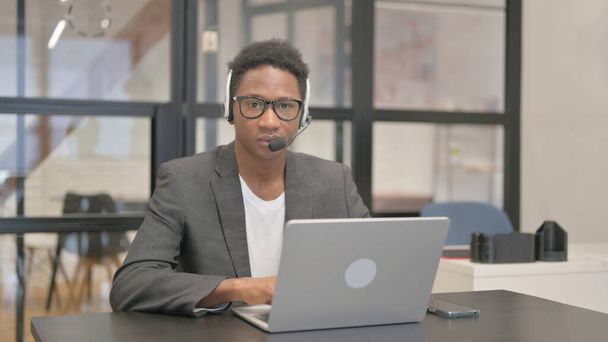 Afroamerikaner mit Headset blickt bei der Arbeit am Laptop in die Kamera - Foto, Bild