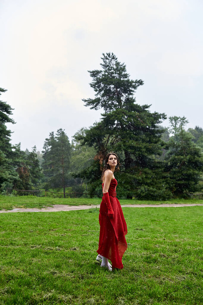 Μια ελκυστική νεαρή γυναίκα σε ένα εντυπωσιακό κόκκινο φόρεμα και μακριά γάντια με χάρη στέκεται σε ένα ηλιόλουστο πεδίο, αισθάνεται το καλοκαιρινό αεράκι. - Φωτογραφία, εικόνα