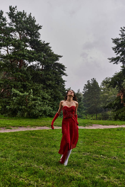 Μια χαριτωμένη νεαρή γυναίκα σε ένα ζωντανό κόκκινο φόρεμα και μακριά γάντια στέκεται μέσα σε καταπράσινο γρασίδι, απολαμβάνοντας το καλοκαιρινό αεράκι. - Φωτογραφία, εικόνα