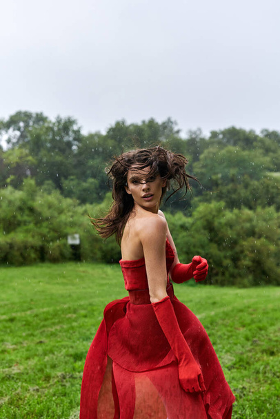 Egy elegáns, vörös ruhás, hosszú kesztyűs fiatal nő áll kecsesen a napfényes mezőn, átázva a nyári szellőben.. - Fotó, kép