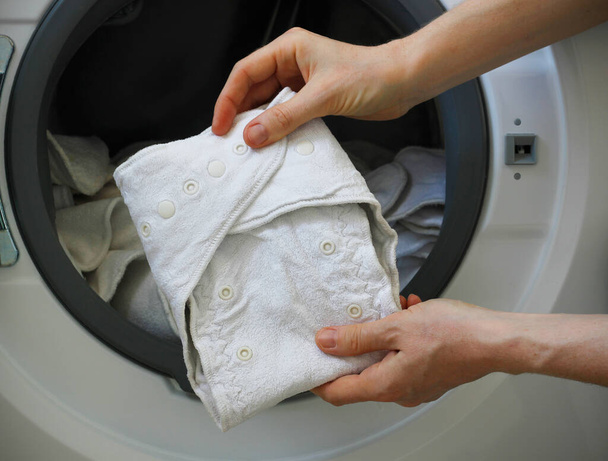 Female hands holding a white cloth baby diaper in front of a washing machine full of cloth bamboo washable diapers. Eco-friendly reusable fabric nappies. Zero waste and sustainability concept - Photo, Image