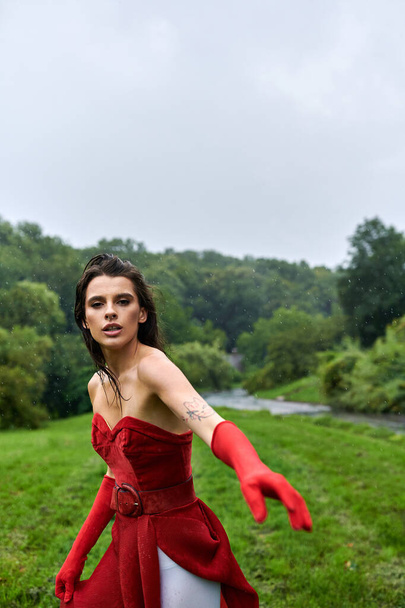Привлекательная молодая женщина в красном платье и длинных перчатках изящно стоит на пышном зеленом поле, наслаждаясь летним бризом. - Фото, изображение