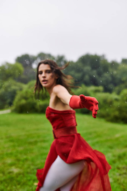 Una joven elegante con un vestido rojo vibrante y guantes largos gira alegremente en medio de la belleza natural de un campo iluminado por el sol. - Foto, Imagen