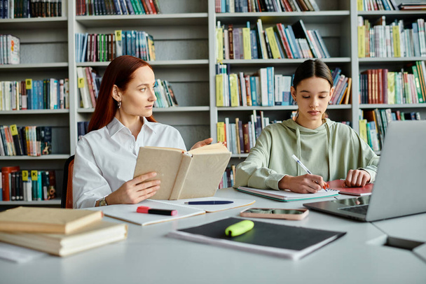 Μια κοκκινομάλλα δασκάλα και μια έφηβη, κάθονται σε ένα τραπέζι βιβλιοθήκης. Ο δάσκαλος διδάσκει ενώ χρησιμοποιεί ένα φορητό υπολογιστή για τη σύγχρονη εκπαίδευση. - Φωτογραφία, εικόνα