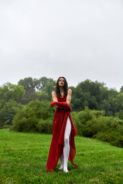 Μια ζωντανή γυναίκα σε ένα κόκκινο φόρεμα και μακριά γάντια με χάρη στέκεται σε ένα πεδίο, βυθισμένο στην απαλή καλοκαιρινή αύρα. - Φωτογραφία, εικόνα