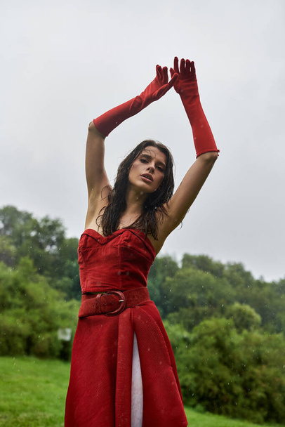 Μια ελκυστική νεαρή γυναίκα σε ένα κόκκινο φόρεμα και μακριά γάντια απολαμβάνοντας το καλοκαιρινό αεράκι στη φύση. - Φωτογραφία, εικόνα