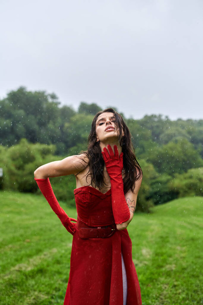 Młoda kobieta w czerwonej sukience i długich rękawiczkach stoi z wdziękiem na polu, ciesząc się letnią bryzą. - Zdjęcie, obraz