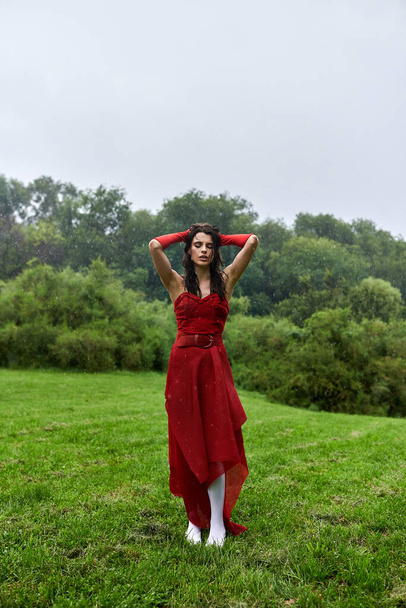 Μια εκπληκτική νεαρή γυναίκα σε ένα ρέον κόκκινο φόρεμα και μακριά γάντια στέκεται με χάρη σε ένα ηλιόλουστο πεδίο, απολαμβάνοντας το καλοκαιρινό αεράκι. - Φωτογραφία, εικόνα