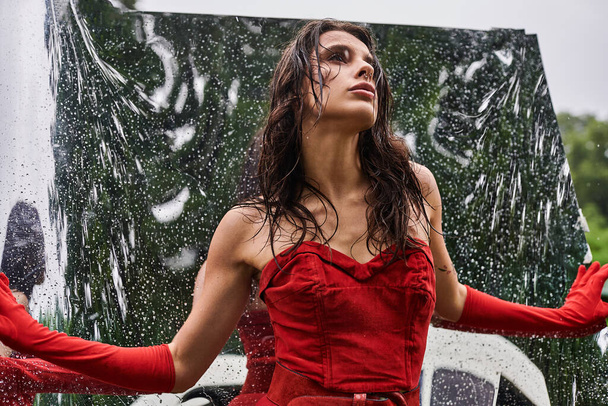 Una joven con un vestido rojo y guantes largos baila con gracia bajo la lluvia, abrazando la refrescante ducha de verano. - Foto, imagen