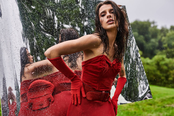 Сяюча жінка в яскраво-червоній сукні витончено стоїть перед дзеркалом, виключаючи елегантність і привабливість. - Фото, зображення