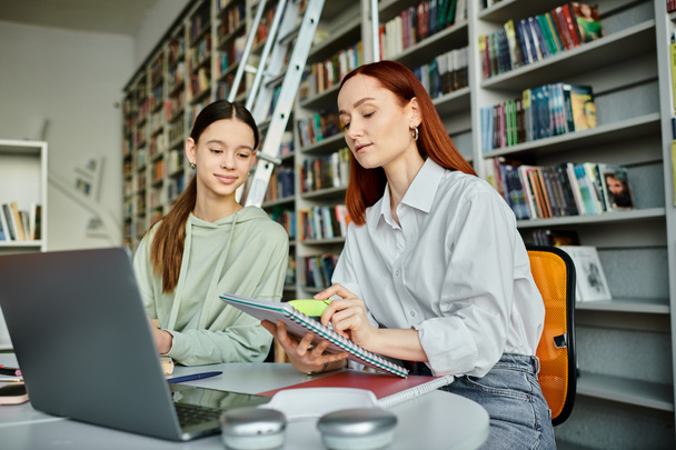 Nachhilfestunde in der Bibliothek, eine rothaarige Tutorin und ein Teenager-Mädchen arbeiten an einem Laptop für moderne Bildung. - Foto, Bild
