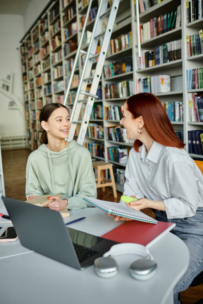 Ein Tutor mit roten Haaren unterrichtet ein Teenager-Mädchen in einer Bibliothek und benutzt einen Laptop für den Nachmittagsunterricht über moderne Bildung. - Foto, Bild