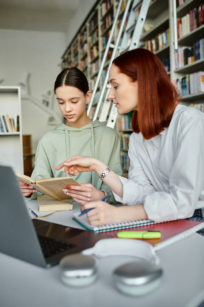 Een tutor met rood haar begeleidt een tiener in een bibliotheek, beide diep gericht op hun laptop schermen voor naschoolse lessen. - Foto, afbeelding