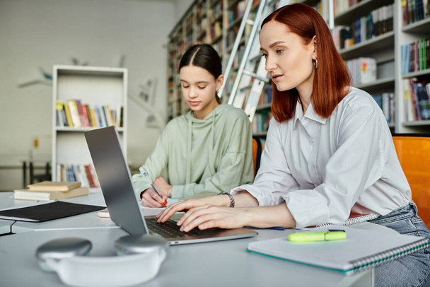 Redhead Tutor engagiert sich nach der Schule Nachhilfestunde mit Teenager-Mädchen in der Bibliothek Einstellung, beide auf Laptop konzentriert. - Foto, Bild
