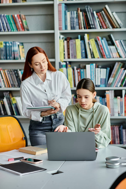 Redhead tutor en tienermeisje deelnemen aan modern onderwijs, bespreken schoollessen op een laptop in een bibliotheek setting. - Foto, afbeelding