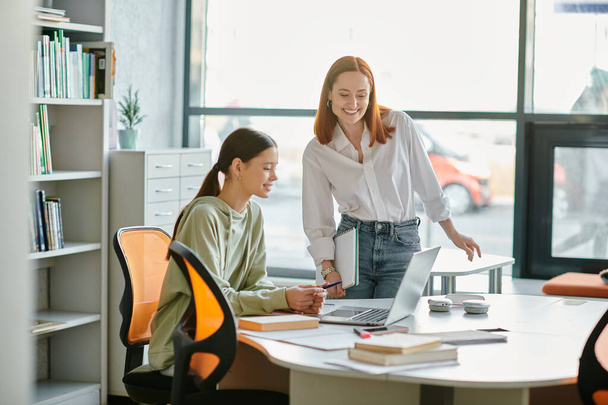 Een roodharige mentor begeleidt een tienerstudent door naschoolse lessen aan een bureau in een modern kantoor, beiden gefascineerd door het laptopscherm.. - Foto, afbeelding