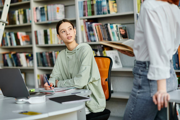 Ein rothaariger Lehrer unterrichtet ein Teenager-Mädchen in einer Bibliothek, beide sitzen an einem Schreibtisch und nutzen einen Laptop für moderne Bildung. - Foto, Bild