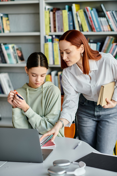 Kızıl saçlı bir öğretmen kütüphanede genç bir öğrenciye ders veriyor. İkisi de bilgisayar ekranına odaklanmış.. - Fotoğraf, Görsel