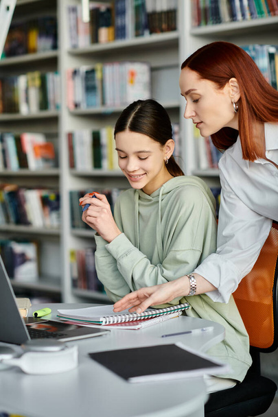 Ένας δάσκαλος με κόκκινα μαλλιά διδάσκει ένα έφηβο κορίτσι σε μια βιβλιοθήκη, καθώς εργάζονται μαζί σε ένα φορητό υπολογιστή για τη σύγχρονη εκπαίδευσή τους. - Φωτογραφία, εικόνα