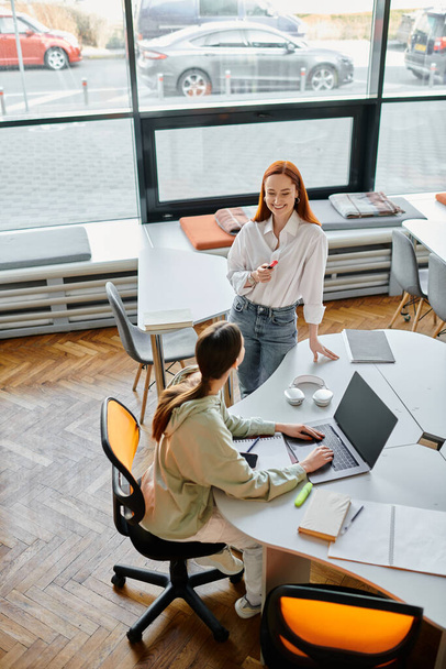 Een roodharige mentor begeleidt een tienermeisje door naschoolse lessen in een moderne kantooromgeving, beiden verdiept in hun werk. - Foto, afbeelding