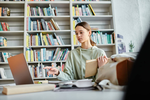 Девушка-подросток сидит за столом, работая над ноутбуком на фоне книжного шкафа в современном образовательном пространстве. - Фото, изображение
