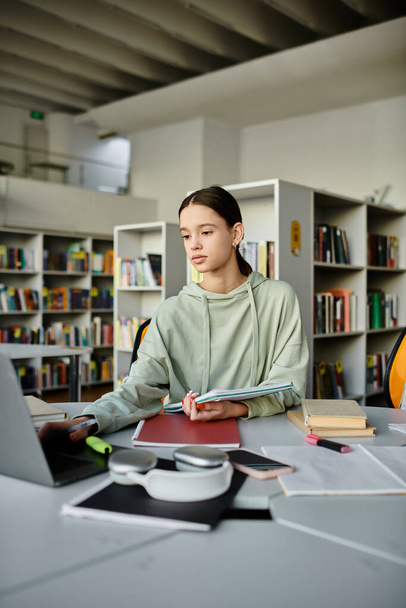 Egy tinédzser lány ül egy íróasztalnál egy könyvtárban, koncentrálva a laptopjára, miközben házi feladatot csinál iskola után.. - Fotó, kép