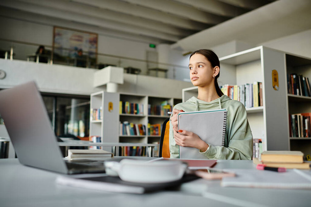 Teini-ikäinen tyttö syvällä ajattelussa, kun hän työskentelee kotitehtävissään kannettavan tietokoneen avulla hiljaisessa kirjastossa. - Valokuva, kuva