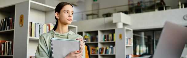 Una adolescente se para frente a una estantería en una biblioteca, inmersa en estudiar y hacer deberes en su computadora portátil después de la escuela. - Foto, imagen