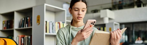 Una ragazza adolescente tiene un libro mentre controlla il suo telefono, immersa nell'educazione moderna e bilanciando i suoi studi con la tecnologia. - Foto, immagini