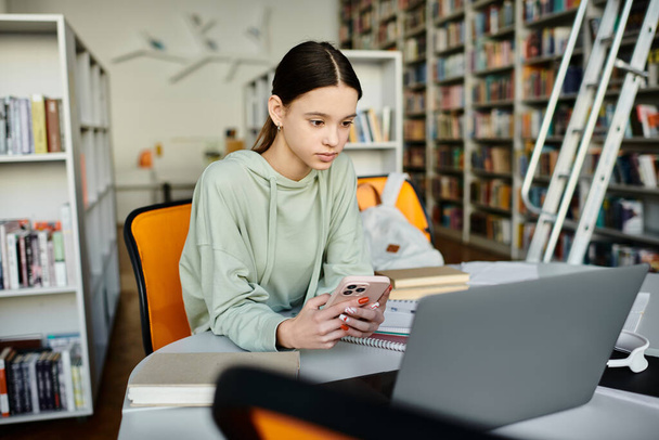 Dospívající dívka sedí za stolem, zaměřuje se na domácí úkoly na svém notebooku a kontroluje, zda její mobil aktualizace o moderním vzdělávání. - Fotografie, Obrázek