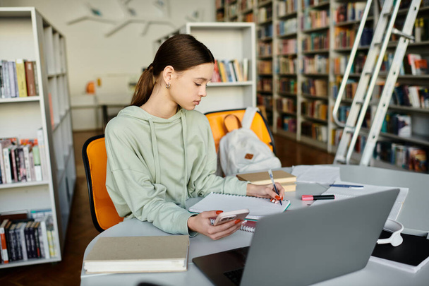 Une adolescente se concentre sur son ordinateur portable à une table dans une bibliothèque, s'engageant dans l'éducation moderne après l'école. - Photo, image