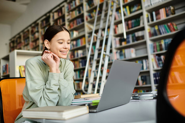 Een tienermeisje zit achter een bureau met een laptop, omringd door boekenplanken, zich intensief bezig houdend met haar huiswerk na school. - Foto, afbeelding