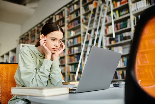 Μια έφηβη με μια ειρηνική έκφραση κάθεται σε ένα γραφείο σε μια βιβλιοθήκη, εστιάζοντας στο laptop της, καθώς κάνει τα μαθήματά της. - Φωτογραφία, εικόνα