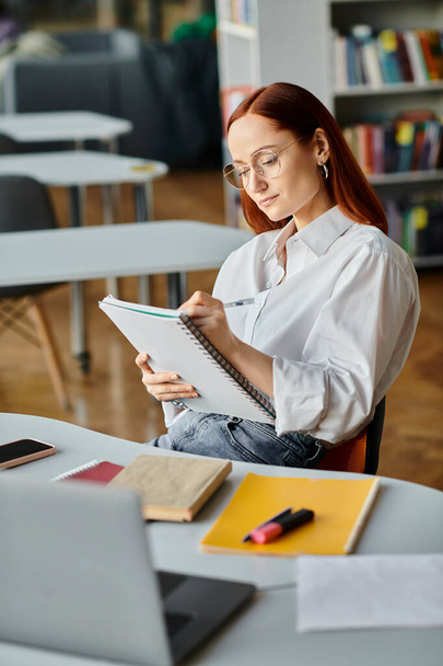 Μια κοκκινομάλλα δασκάλα κάθεται στο γραφείο της, επικεντρωμένη στο γράψιμο σε ένα σημειωματάριο μετά από ένα διαδικτυακό μάθημα.. - Φωτογραφία, εικόνα
