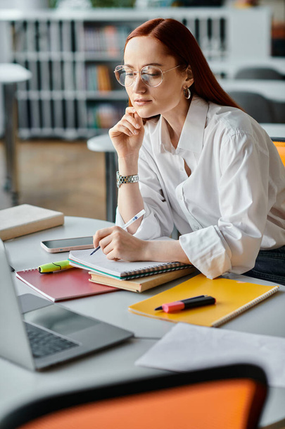 Egy vörös hajú tanítónő ül egy asztalnál, aki egy iskolai lecke után laptoppal tanít az interneten.. - Fotó, kép