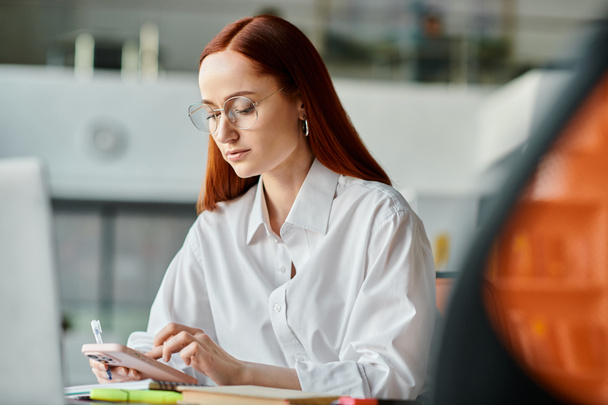 Egy vörös hajú női oktató online tanít, asztalnál ül és iskola után kapcsolatba lép egy okostelefonnal.. - Fotó, kép
