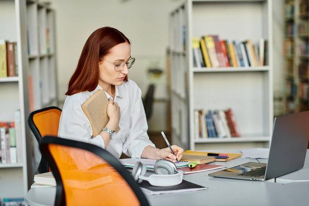 Μια γυναίκα καθηγήτρια με κόκκινα μαλλιά που παρέχει ένα μάθημα μετά το σχολείο στο διαδίκτυο, χρησιμοποιώντας ένα φορητό υπολογιστή σε ένα γραφείο σε μια ρύθμιση βιβλιοθήκης. - Φωτογραφία, εικόνα