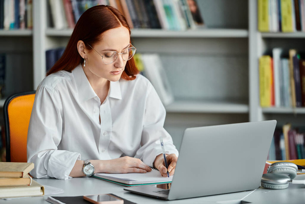 A vörös hajú női oktató összpontosít tanítani egy iskola utáni leckét, miközben dolgozik egy laptop a könyvtárban. - Fotó, kép
