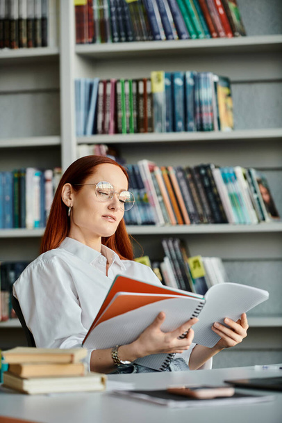 Een roodharige vrouw verdiept in een boek in een bibliotheek, omarmd door de eenzaamheid van haar lezen. - Foto, afbeelding