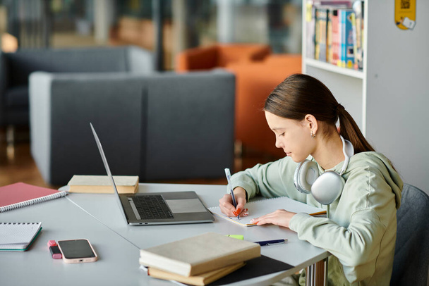 egy tinédzser lány egy asztalnál, aki laptoppal van elfoglalva, miközben házi feladatot csinál. - Fotó, kép