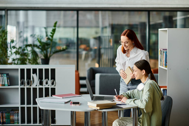 Een roodharige vrouw begeleidt een tienermeisje aan een bureau in een modern kantoor, beiden verdiept in hun studie met een laptop. - Foto, afbeelding