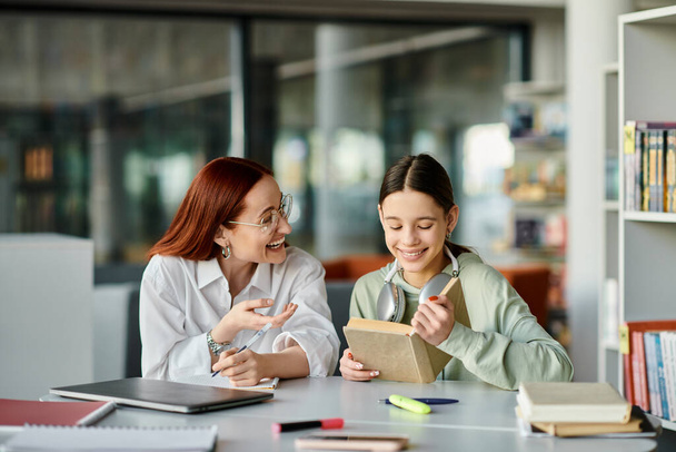 Una rossa insegna a un'adolescente in una biblioteca, assorta in lezioni post-scolastiche con un computer portatile. - Foto, immagini