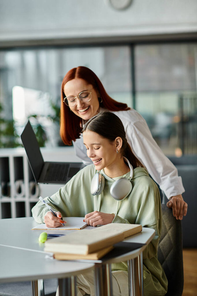 Μια κοκκινομάλλα διδάσκει ένα έφηβο κορίτσι σε ένα τραπέζι σε ένα γραφείο, και οι δύο επικεντρώθηκαν σε ένα φορητό υπολογιστή κατά τη διάρκεια των μαθημάτων μετά το σχολείο. - Φωτογραφία, εικόνα