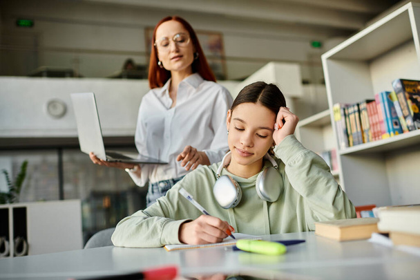 Un tutor - una donna rossa - sta insegnando a una ragazza adolescente a una scrivania usando un computer portatile per le lezioni post-scolastiche. - Foto, immagini