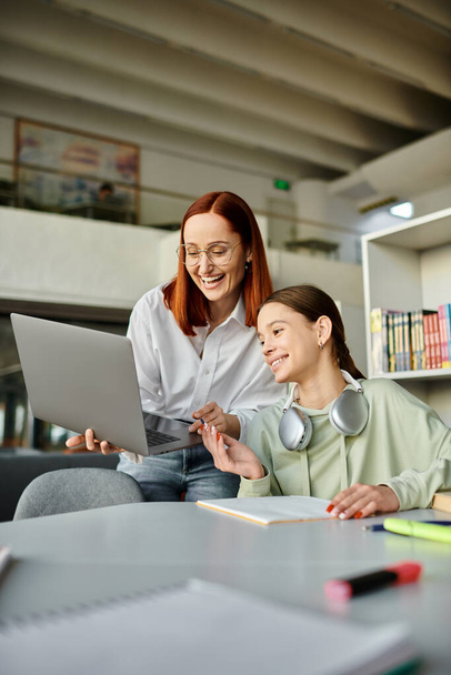 Mujer pelirroja tutores una adolescente en una biblioteca, ambos centrados en un ordenador portátil, participan en lecciones después de la escuela. - Foto, imagen