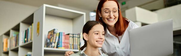 Una donna dai capelli rossi tiene un portatile davanti a una libreria mentre insegna a una ragazza adolescente, ridefinendo l'educazione moderna.. - Foto, immagini