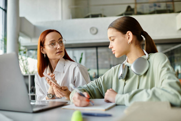Een roodharige vrouw begeleidt een tienermeisje aan een bureau, bezig met een gesprek terwijl het gebruik van een laptop voor modern onderwijs. - Foto, afbeelding