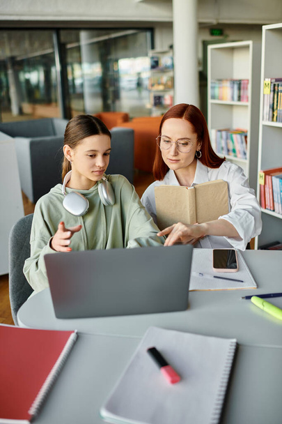 Femme rousse enseignant adolescente, tuteur et élève ayant des leçons après l'école tout en travaillant sur un ordinateur portable dans une bibliothèque. - Photo, image